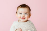 Joyful baby wearing cozy white knit sweater laughs and smiles against soft pink background, capturing pure happiness and innocence in childhood portrait.