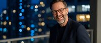 Confident mature businessman with glasses smiling at camera against blurred city lights background. Professional corporate executive portrait for business success concepts.
