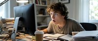 Young man with curly brown hair and light complexion, wearing casual beige shirt, focused on computer monitor at desk with coffee cup and papers, modern home office setting with shelving.