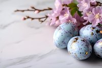 Decorative blue speckled Easter eggs with gold flakes and delicate pink cherry blossoms on white marble background, creating elegant spring holiday composition.