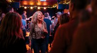 Mature blonde woman dancing and socializing at lively evening party with string lights and crowd of people enjoying nightlife entertainment and celebration.