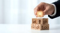 Businessman hand placing wooden cube with lightbulb icon on stack of business symbol blocks representing innovation and strategic planning concepts.