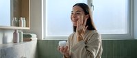Young woman applying skincare cream in modern bathroom with green tiles and natural light for daily beauty routine.
