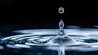 Mesmerizing water drop collision captured in high speed photography creating crown splash formation with concentric ripples on dark blue surface. Perfect for wellness.