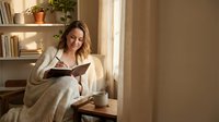 Young woman reading book in cozy armchair by window with warm natural light, relaxing at home with coffee cup and plants creating peaceful atmosphere.