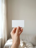 Hand holding blank white business card against neutral beige wall with natural window light, suitable for branding, corporate identity, and mockup designs.