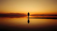 Silhouette of person standing on reflective beach at golden sunset with mirror reflection in wet sand creating peaceful meditation scene.
