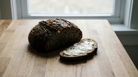 Freshly baked dark rye bread loaf with sliced piece on wooden table near window, showcasing crusty exterior and soft interior for bakery and food styling.