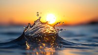 Dynamic water splash wave catching golden sunlight during dramatic ocean sunset, with droplets frozen in motion against glowing orange sky and dark blue waters.