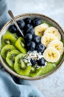 Green smoothie bowl topped with fresh kiwi slices, blueberries, banana, and coconut flakes served in rustic ceramic bowl. Healthy breakfast concept.