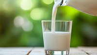 Fresh milk pouring from bottle into clear glass on wooden table with soft green bokeh background, morning breakfast concept for healthy lifestyle.