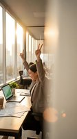 Successful Asian businesswoman celebrating victory at office desk with laptop showing growth charts, raising arms in triumph by sunny windows.