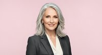 Mature woman with gray wavy hair, fair complexion, wearing black blazer and white blouse against pink background. Professional business portrait.