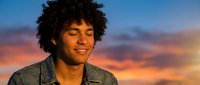 Young mixed-race man with curly hair smiling peacefully with eyes closed against vibrant sunset sky. Relaxation and mindfulness concept for wellness content.