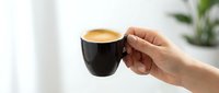 Person holding black coffee cup with latte art, enjoying hot beverage in bright interior space with plant in background.