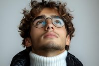 Young man with curly hair and glasses looking up thoughtfully, wearing white turtleneck sweater and leather jacket against light background.