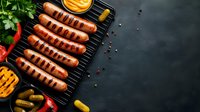 Grilled sausages on black cast iron grill pan with mustard sauce, pickles, red chili peppers and fresh herbs on dark background with copy space.