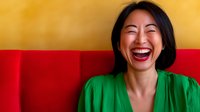 Young Asian woman with genuine laughter wearing green blouse against vibrant yellow and red background, expressing pure joy and happiness in casual portrait setting.