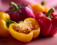 Fresh colorful bell peppers in red, yellow and purple varieties with one cut open showing seeds and inner texture on pink background, macro food photography.