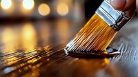 Paint brush applying brown wood stain or varnish on wooden surface with golden bokeh lights in background, macro photography showing detail of bristles.