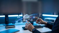 Young male professional with dark skin tone working at modern desk with multiple monitors, interacting with holographic cloud computing interface and digital network visualization for IT, software development, or data management projects.