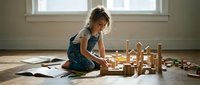 Young girl playing with wooden building blocks on floor creating imaginative structures in bright sunlit room with scattered papers and toys around.