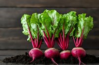 Fresh organic beetroots with vibrant green leaves lined up on dark soil against rustic wooden background, showcasing farm-to-table produce and natural gardening.