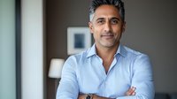 Middle-aged South Asian businessman with confident smile wearing blue striped shirt, standing with arms crossed in modern office setting. Natural window lighting.