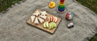 Overhead view of sliced cheese and apple on wooden board with colorful toys on picnic blanket, ideal for family outdoor dining and children's food content.