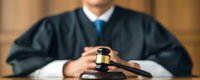 Male judge in traditional black robe and blue tie sitting at courtroom bench with wooden gavel, representing justice system and legal proceedings in professional setting.