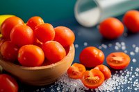 Fresh cherry tomatoes in wooden bowl with sea salt scattered on dark background, some cut in half showing juicy texture, close-up food photography.