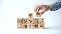 Hand placing wooden blocks with educational icons including books, gears, plants and learning symbols on white background for skill development concepts.