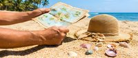 Man with sunburned skin holding map on sandy beach with straw hat and seashells, planning summer vacation travel destination by ocean.