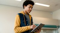 Young male student of mixed ethnicity wearing yellow long-sleeve shirt and denim vest, holding tablet device in classroom setting, engaged in digital learning and education.