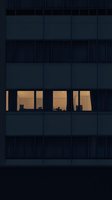 Illuminated office windows in dark modern building facade reveal silhouettes of desks and furniture, creating dramatic contrast between warm interior and cool exterior.