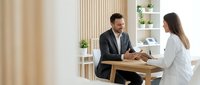 Male professional in dark blazer and female professional in white shirt having business discussion at wooden desk in modern office setting with neutral decor.