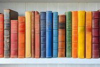 Row of vintage hardcover books in various colors arranged on white wooden shelf, showing spines with classic binding and embossed titles. Library collection display.