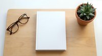 Minimalist workspace with blank white book, eyeglasses and succulent plant on wooden desk. Clean office setup for reading, writing or creative projects.