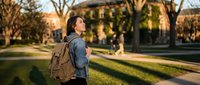 Young Asian woman student with backpack walking on university campus during golden hour sunset, looking up with hopeful expression for education concept.