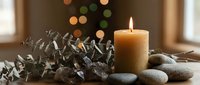 Burning candle surrounded by smooth stones and eucalyptus branches creates peaceful spa atmosphere with warm bokeh lights in cozy interior setting.