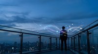 Young Asian businessman standing on modern glass observation deck overlooking city skyline at dusk with digital network connections and data visualization.