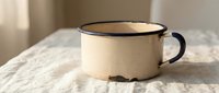 Vintage enamel mug with dark rim sits on white linen fabric in soft natural light creating cozy morning atmosphere for kitchen and home decor themes.