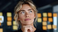 Young blonde businesswoman looking up thoughtfully with hand on chin in modern office environment with warm lighting and blurred background.