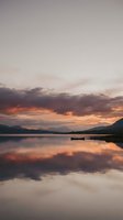 Serene lake landscape at sunset with dramatic orange and purple clouds reflected in calm water, mountains in distance, small boat moored nearby.