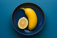 Bright yellow lemon shaped like a banana on a blue ceramic plate, bold color contrast, creative food concept on teal background.