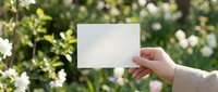 Hand holding blank white card mockup outdoors in spring garden with blooming flowers, green bokeh background, copy space for text.