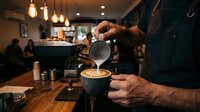 Barista pouring steamed milk creating latte art in coffee cup at modern cafe with warm lighting and customers in background for hospitality business.
