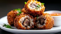 Deep fried rice balls filled with tender meat and melted cheese, garnished with fresh herbs and served with dipping sauce on white plate against dark background.