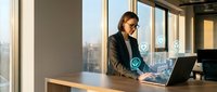 Young adult male with dark hair and glasses, wearing a dark blazer, working at a laptop in a modern high-rise office with floor-to-ceiling windows overlooking a city skyline, suitable for business and corporate concepts.