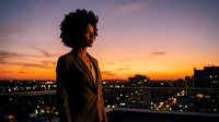 Young African American businesswoman in suit standing on urban rooftop at sunset with city lights, professional lifestyle and career success concept.